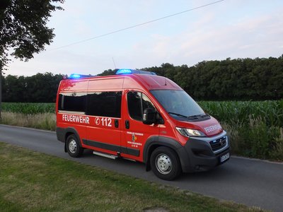 Mannschaftstransportwagen des Löschzugs St. Vit steht auf einem kleinen Weg mit eingeschaltetem Blaulicht. Im Vordergrund befindet sich eine Wiese und im Hintergrund ein Meisfeld.